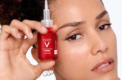 Woman smiling whilst holding up two red coloured Vichy Liftactiv B3 products: the serum and the day cream.
